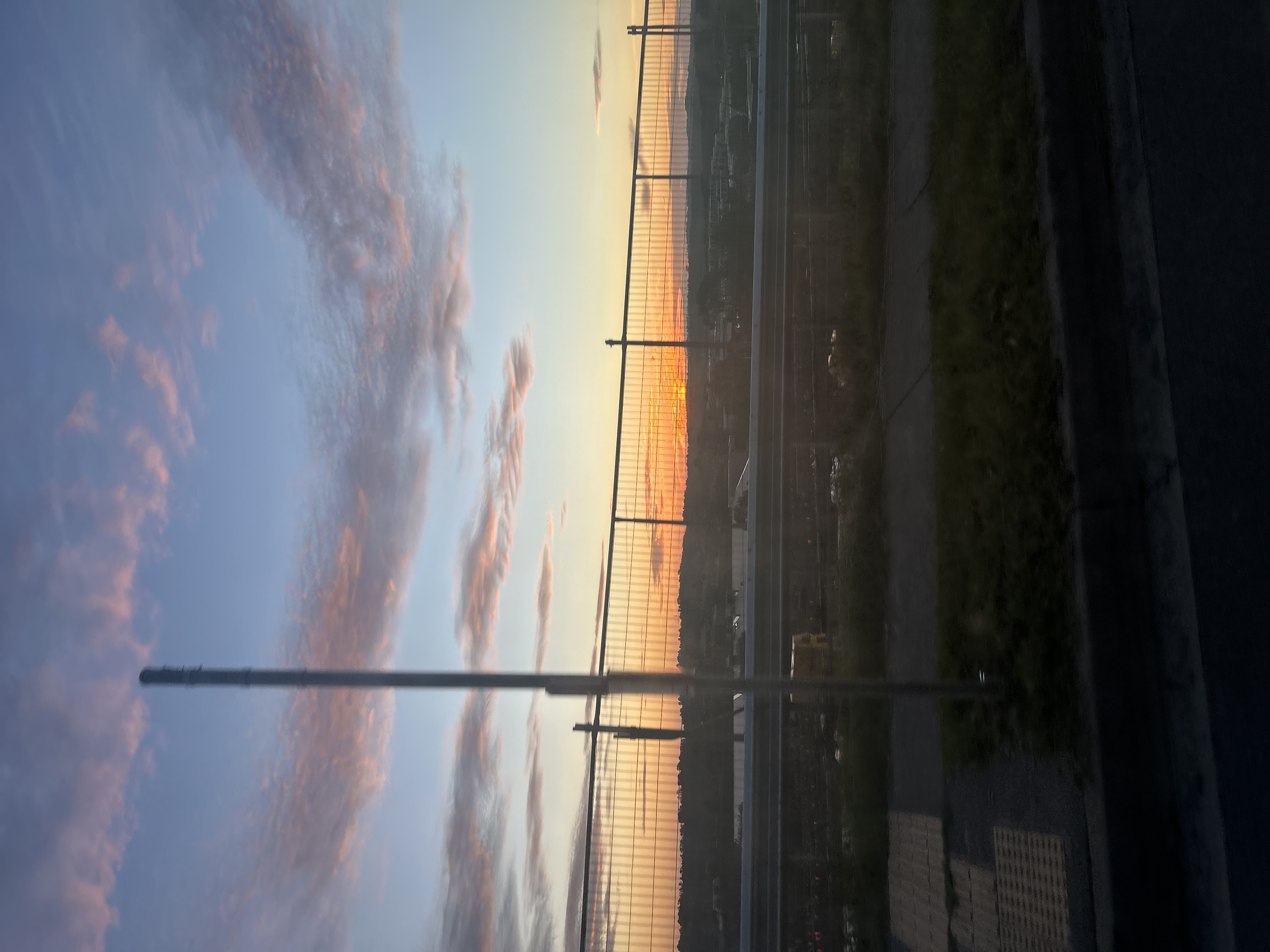 Colorful sunset sky with soft clouds above a roadway and metal fence. Sky transitions from light blue at the top to pale yellow and orange near the horizon, with streaks of pink and purple clouds scattered across it.