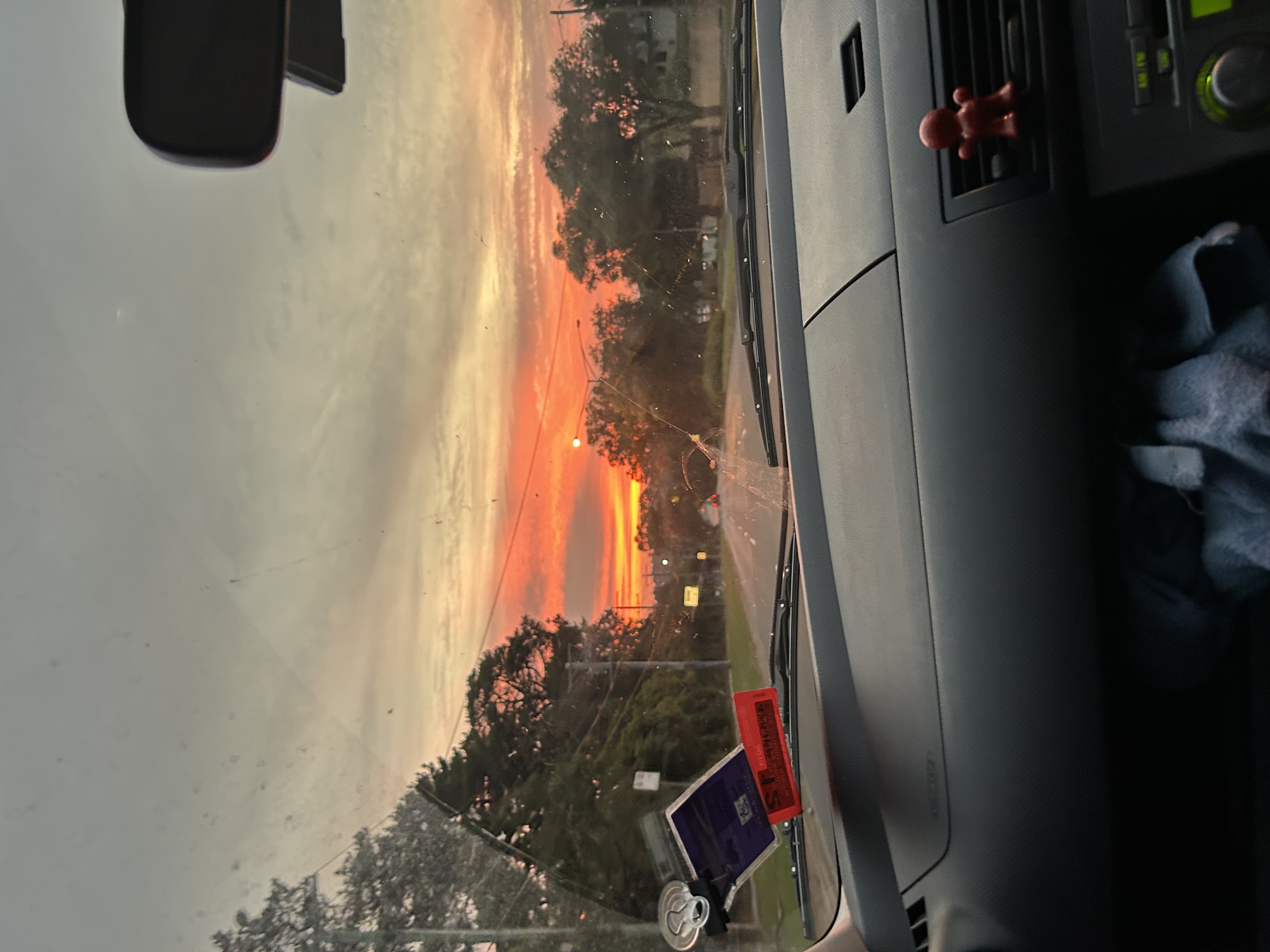 Sunset sky in shades of orange, red, and pink stretching across the horizon ahead of a car on a road.Dark tree silhouettes line both sides of the road and frame the bright strip of sunset low in the center distance. The road itself is mostly empty, with just a few faint car lights far ahead.
