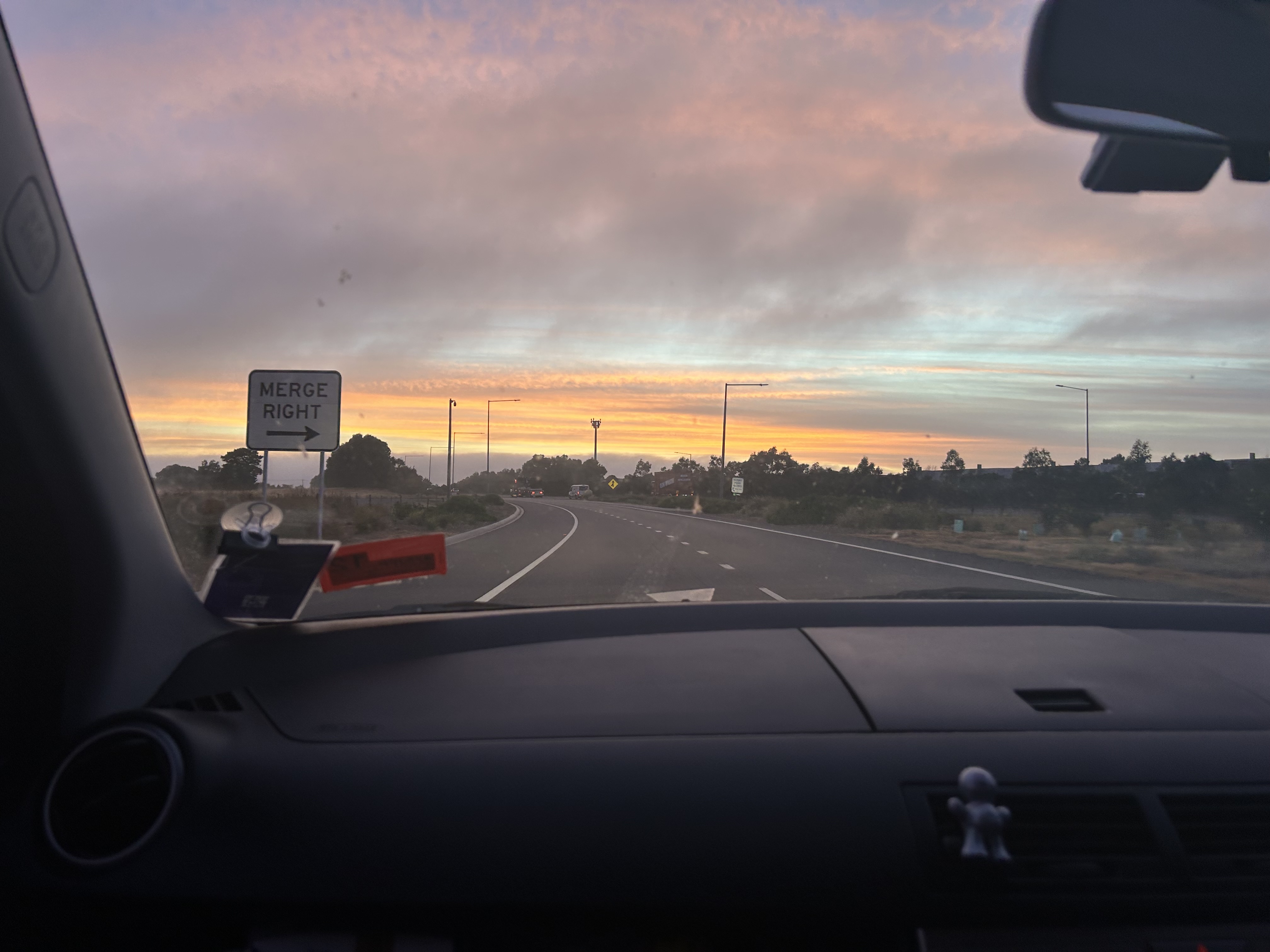 The image is taken from the front passenger seat of a car driving on a divided highway. In the middle distance, a white road sign on the left side reads “MERGE RIGHT” with a right-pointing arrow. The road curves gently to the right, with dashed lane markings and several tall, thin streetlights along the sides. A few cars are visible far ahead. Low bushes and trees line the road, and the sky is filled with layered clouds colored pink, orange, and blue from the low sun near the horizon.